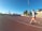 A person is jogging on a red athletics track under a clear blue sky. They are wearing a light gray top with orange shorts and running shoes. The track surrounds a field with sparse trees, and there is a set of bleachers to the right. A tall lamppost is visible, suggesting it is a sports facility.