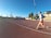 A person is jogging on a red athletics track under a clear blue sky. They are wearing a light gray top with orange shorts and running shoes. The track surrounds a field with sparse trees, and there is a set of bleachers to the right. A tall lamppost is visible, suggesting it is a sports facility.