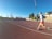 A person is jogging on a red athletics track under a clear blue sky. They are wearing a light gray top with orange shorts and running shoes. The track surrounds a field with sparse trees, and there is a set of bleachers to the right. A tall lamppost is visible, suggesting it is a sports facility.