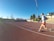 A person is jogging on a red athletics track under a clear blue sky. They are wearing a light gray top with orange shorts and running shoes. The track surrounds a field with sparse trees, and there is a set of bleachers to the right. A tall lamppost is visible, suggesting it is a sports facility.