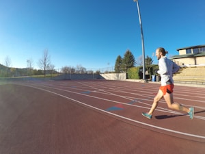 A person is jogging on a red athletics track under a clear blue sky. They are wearing a light gray top with orange shorts and running shoes. The track surrounds a field with sparse trees, and there is a set of bleachers to the right. A tall lamppost is visible, suggesting it is a sports facility.