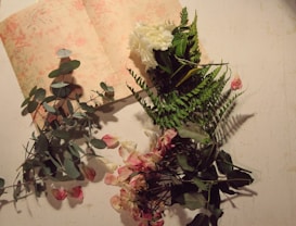 An arrangement of dried leaves and flowers, including eucalyptus, ferns, and pink roses, is spread across an open book with floral patterns on its pages.