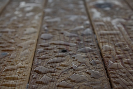 Drops of water scattered across a wooden surface, highlighting the wood grain texture. The surface appears wet with varying sizes of water droplets.