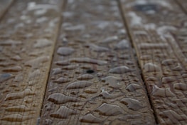 Drops of water scattered across a wooden surface, highlighting the wood grain texture. The surface appears wet with varying sizes of water droplets.