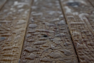 Drops of water scattered across a wooden surface, highlighting the wood grain texture. The surface appears wet with varying sizes of water droplets.