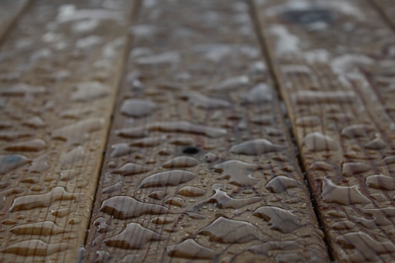 Drops of water scattered across a wooden surface, highlighting the wood grain texture. The surface appears wet with varying sizes of water droplets.
