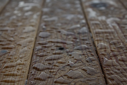 Drops of water scattered across a wooden surface, highlighting the wood grain texture. The surface appears wet with varying sizes of water droplets.