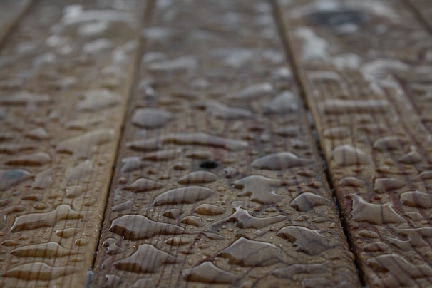 Drops of water scattered across a wooden surface, highlighting the wood grain texture. The surface appears wet with varying sizes of water droplets.