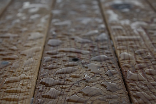 Drops of water scattered across a wooden surface, highlighting the wood grain texture. The surface appears wet with varying sizes of water droplets.