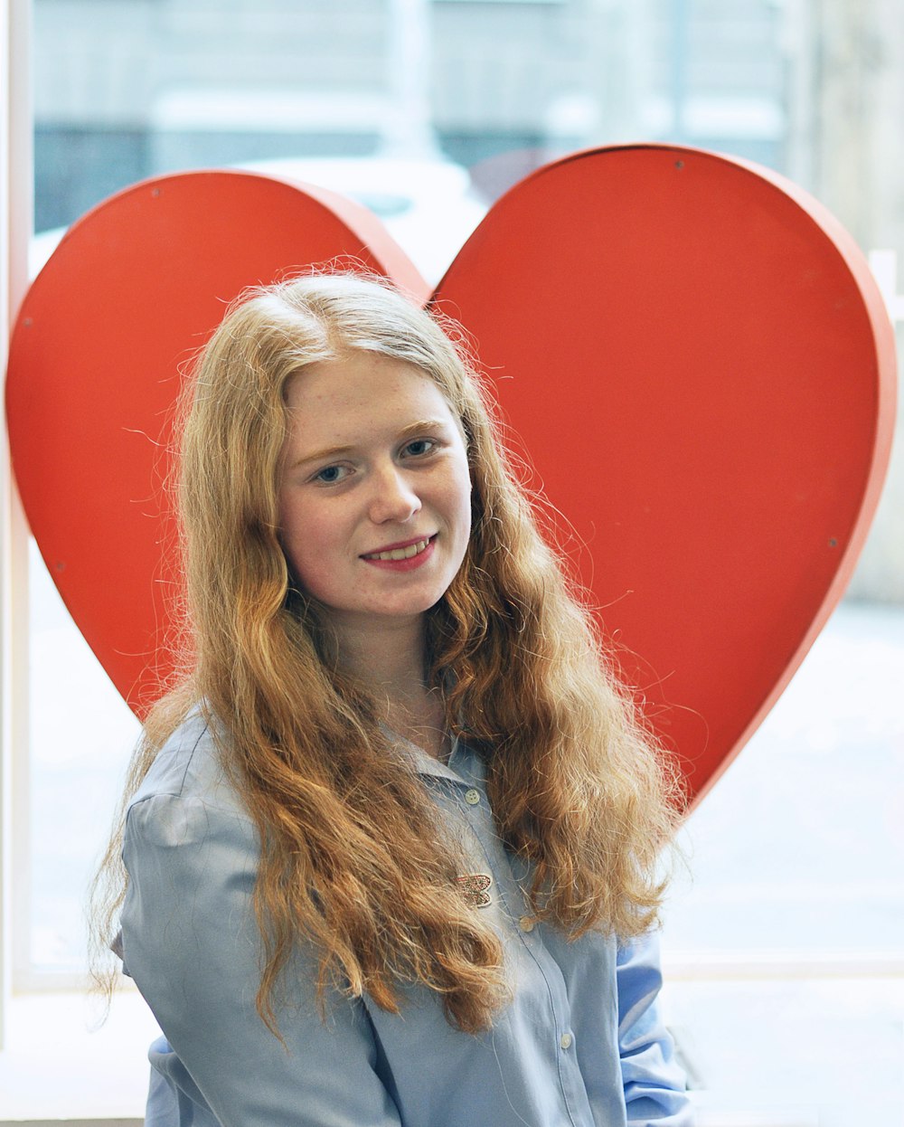 Una mujer rubia de pie frente a un gran corazón rojo.