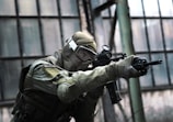 A person in tactical military gear holding a rifle and aiming intently. The gear includes a helmet, protective eyewear, and a camouflaged uniform. The backdrop consists of large industrial windows, suggesting an urban or abandoned setting.