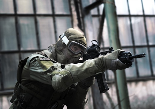 A person in tactical military gear holding a rifle and aiming intently. The gear includes a helmet, protective eyewear, and a camouflaged uniform. The backdrop consists of large industrial windows, suggesting an urban or abandoned setting.