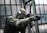 A person in tactical military gear holding a rifle and aiming intently. The gear includes a helmet, protective eyewear, and a camouflaged uniform. The backdrop consists of large industrial windows, suggesting an urban or abandoned setting.