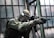 A person in tactical military gear holding a rifle and aiming intently. The gear includes a helmet, protective eyewear, and a camouflaged uniform. The backdrop consists of large industrial windows, suggesting an urban or abandoned setting.