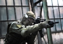 A person in tactical military gear holding a rifle and aiming intently. The gear includes a helmet, protective eyewear, and a camouflaged uniform. The backdrop consists of large industrial windows, suggesting an urban or abandoned setting.