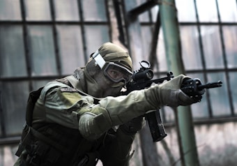 A person in tactical military gear holding a rifle and aiming intently. The gear includes a helmet, protective eyewear, and a camouflaged uniform. The backdrop consists of large industrial windows, suggesting an urban or abandoned setting.
