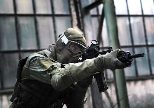 A person in tactical military gear holding a rifle and aiming intently. The gear includes a helmet, protective eyewear, and a camouflaged uniform. The backdrop consists of large industrial windows, suggesting an urban or abandoned setting.