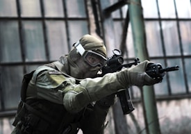 A person in tactical military gear holding a rifle and aiming intently. The gear includes a helmet, protective eyewear, and a camouflaged uniform. The backdrop consists of large industrial windows, suggesting an urban or abandoned setting.