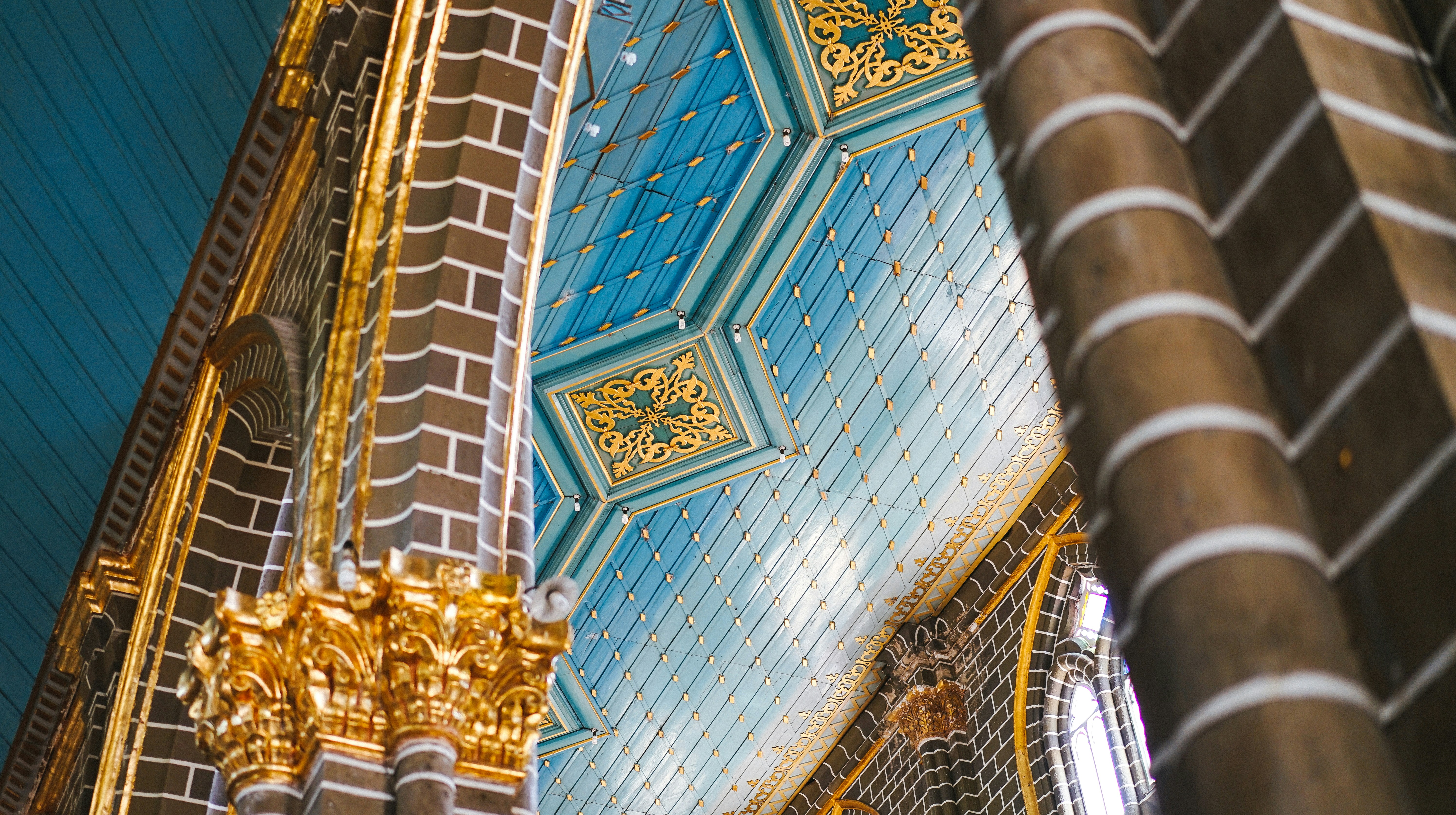The intricate interior of a building features ornate architectural details. Golden decorative elements adorn richly textured brown columns. The ceiling is painted blue, embellished with gold patterns and accents, adding a touch of elegance and grandeur to the setting.