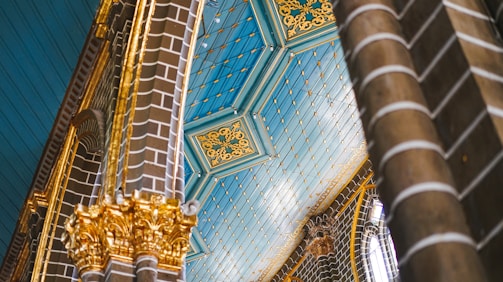 The intricate interior of a building features ornate architectural details. Golden decorative elements adorn richly textured brown columns. The ceiling is painted blue, embellished with gold patterns and accents, adding a touch of elegance and grandeur to the setting.