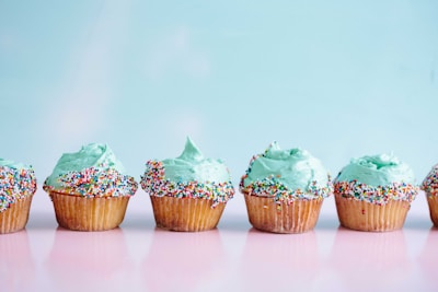 Five cupcakes with light blue frosting are arranged in a row on a reflective pink surface. Each cupcake is decorated with colorful sprinkles, adding a playful contrast to the pastel background.