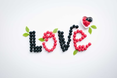The word 'love' is spelled out using black and red gummy candies shaped like berries, with green candy leaves interspersed. A small white heart-shaped dish filled with additional gummy candies is placed near the word.