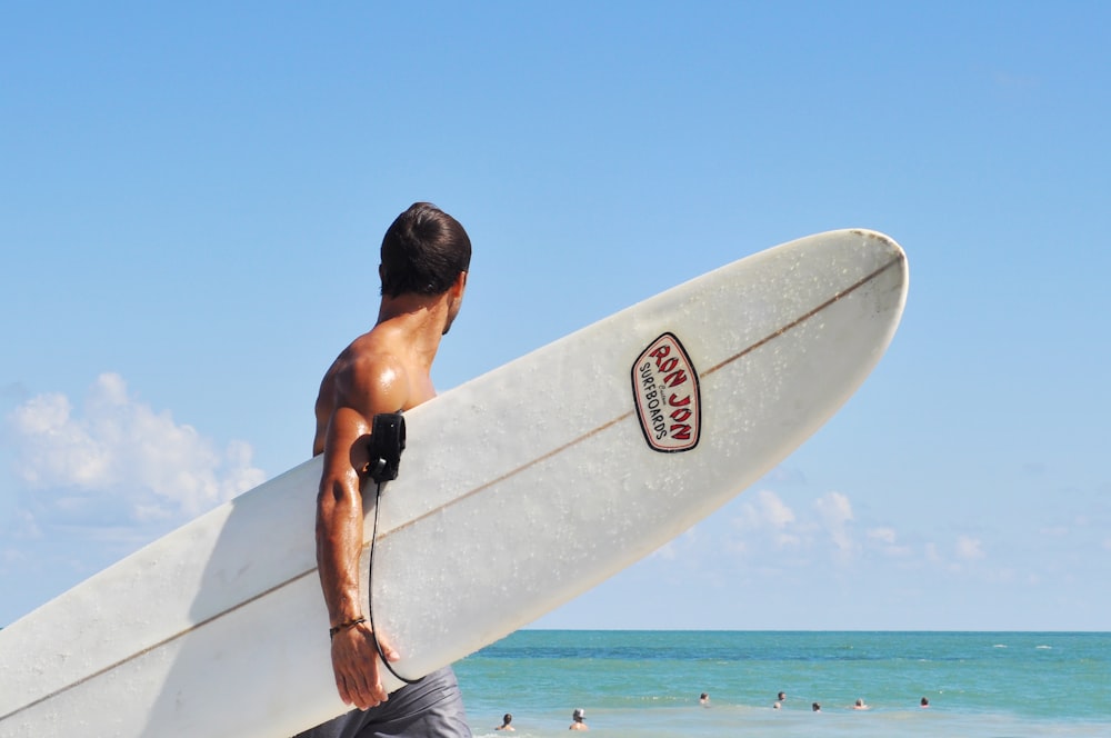 uomo in topless che trasporta tavola da surf bianca in spiaggia durante il giorno