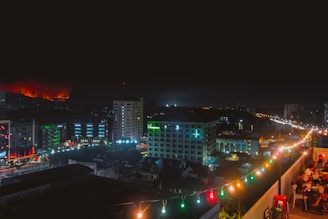 The city skyline at night viewed from the rooftop, framed by neon-lit shisha pipes and lively guests.