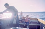 man holding gray fish standing beside two boys sitting on boat seats