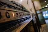 A vintage elevator panel featuring a sequence of numbers with a highlighted number one in orange. The numbers are embedded in a wooden panel with geometric designs, set in an industrial or historical building interior.