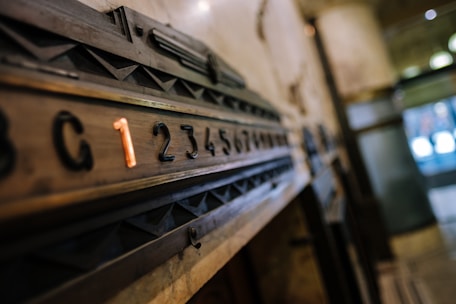 A vintage elevator panel featuring a sequence of numbers with a highlighted number one in orange. The numbers are embedded in a wooden panel with geometric designs, set in an industrial or historical building interior.