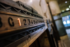 A vintage elevator panel featuring a sequence of numbers with a highlighted number one in orange. The numbers are embedded in a wooden panel with geometric designs, set in an industrial or historical building interior.