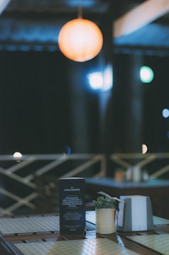 A warmly lit outdoor setting at night with a focus on a table. The table houses a small plant, a menu or flyer with text, and a condiment holder. Blurred lights in the background suggest a cozy, intimate ambiance.