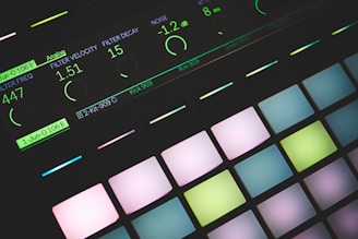 A close-up view of a music production device featuring illuminated pads and a digital display showing various sound parameters. The pads are backlit with pastel colors, and the display indicates settings like filter velocity, decay, and noise levels.