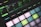 A close-up view of a music production device featuring illuminated pads and a digital display showing various sound parameters. The pads are backlit with pastel colors, and the display indicates settings like filter velocity, decay, and noise levels.