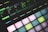 A close-up view of a music production device featuring illuminated pads and a digital display showing various sound parameters. The pads are backlit with pastel colors, and the display indicates settings like filter velocity, decay, and noise levels.