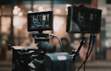A professional video camera setup is present, with two screens displaying the footage being recorded. The background is blurred, showing warm lights and an indistinct environment.