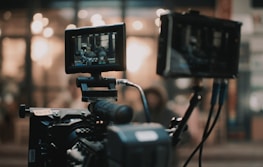 A professional video camera setup is present, with two screens displaying the footage being recorded. The background is blurred, showing warm lights and an indistinct environment.