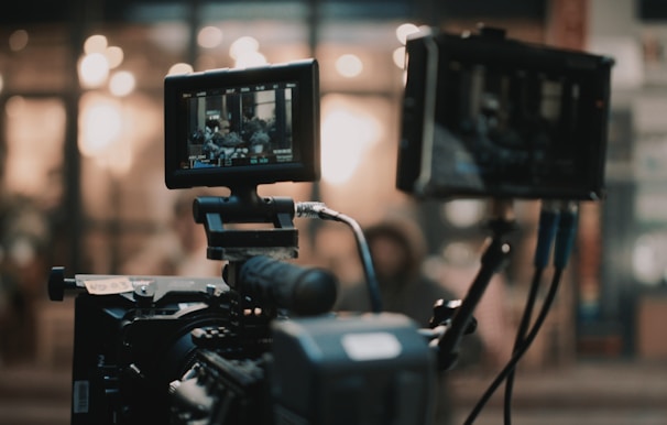 A professional video camera setup is present, with two screens displaying the footage being recorded. The background is blurred, showing warm lights and an indistinct environment.