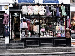 A storefront with a black facade displays various colorful vintage clothing items. Dresses of different styles and patterns are hung outside on hangers and mannequins. Inside the large glass window, more clothing and accessories are visible, along with signs advertising sales and leasing information. The shop entrance, marked with the number 55, appears on the left side.