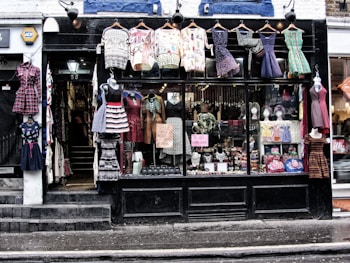 A storefront with a black facade displays various colorful vintage clothing items. Dresses of different styles and patterns are hung outside on hangers and mannequins. Inside the large glass window, more clothing and accessories are visible, along with signs advertising sales and leasing information. The shop entrance, marked with the number 55, appears on the left side.