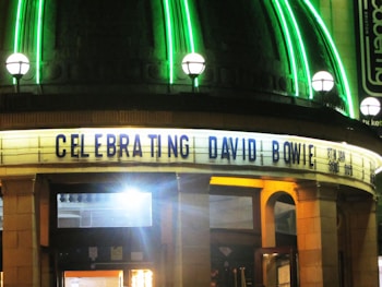 A theater marquee brightly lit at night with neon green lights above, displaying the text 'CELEBRATING DAVID BOWIE 8TH JAN SOLD OUT' in bold letters. The structure has an art-deco style with arched doorways illuminated by warm yellow lighting.