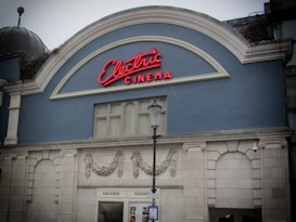 A vintage cinema building facade featuring a pastel blue wall and white decorative arches. A neon sign reading 'Electric Cinema' in red is mounted above, and a street lamp is positioned in front of the entrance.