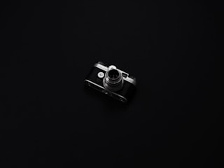A vintage camera with metallic and black elements is centered on a minimalist, dark background. The camera's intricate details, such as the lens and dials, are highlighted by the low lighting, creating a sense of nostalgia and elegance.