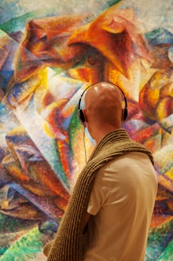 A person wearing headphones stands in front of an abstract, colorful painting. The painting features vibrant swirls and geometric shapes, creating a dynamic composition. The viewer is looking intently at the art, with a knit scarf draped over their shoulders.