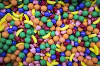 A colorful assortment of various shaped candies including round, spherical, and banana-shaped sweets. The candies display a variety of textures, with some appearing smooth and others with a bumpy surface.