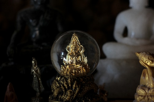 A golden statue enclosed in a clear glass sphere is surrounded by various other figurines. The statues include both seated and standing figures, with one prominent white statue in the background.
