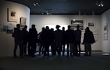 A group of people stands closely together in front of a well-lit display in an art gallery, observing the artwork. The gallery walls are adorned with several framed pieces, creating a contemplative atmosphere.