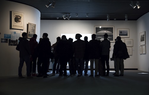 A group of people stands closely together in front of a well-lit display in an art gallery, observing the artwork. The gallery walls are adorned with several framed pieces, creating a contemplative atmosphere.
