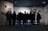 A group of people stands closely together in front of a well-lit display in an art gallery, observing the artwork. The gallery walls are adorned with several framed pieces, creating a contemplative atmosphere.